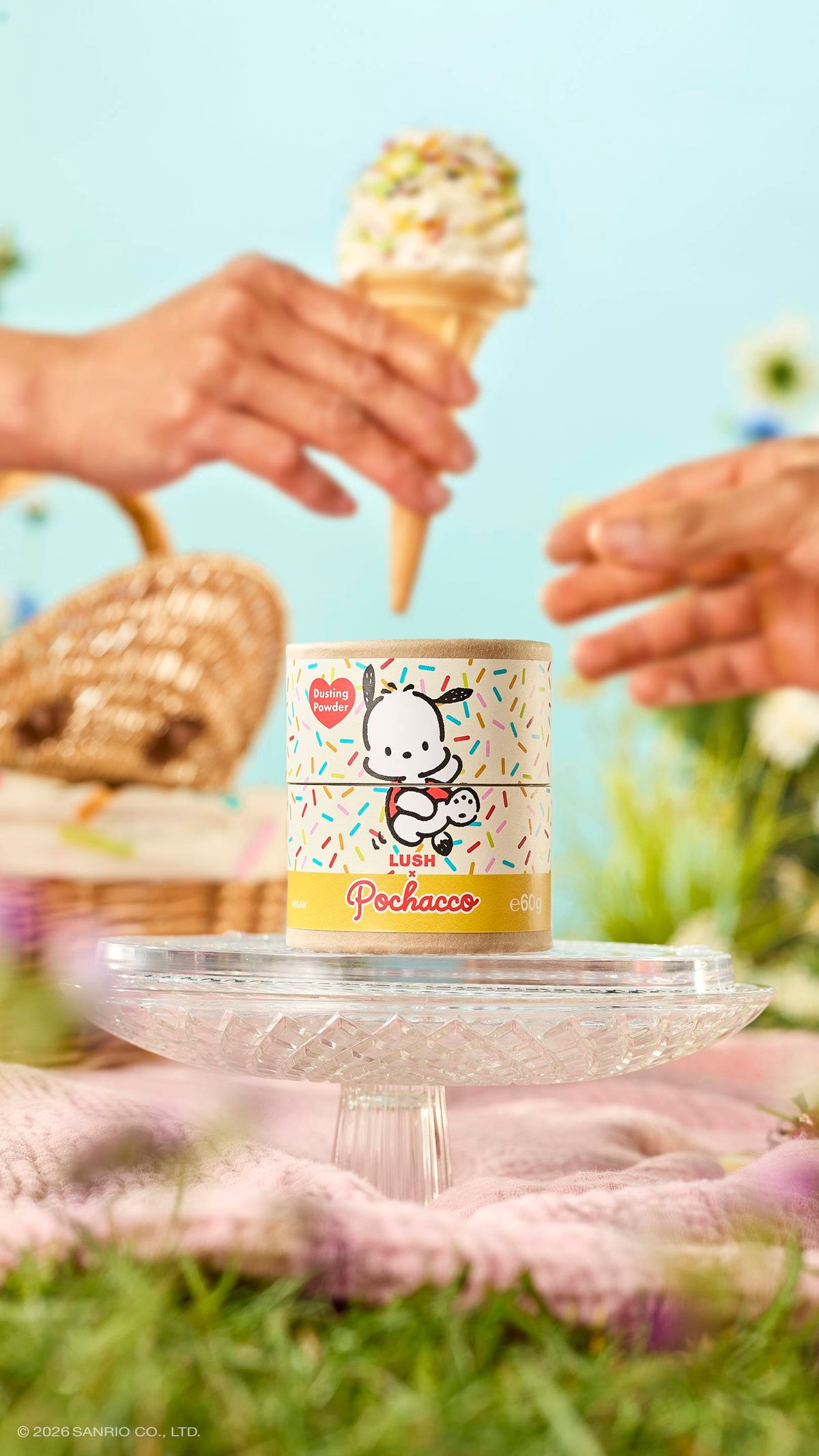 A cardboard tube of Lush x Pochacco dusting powder sits on a glass stand in a meadow. Above it, one hand passes a sprinkle-topped ice cream cone to another. The sunny picnic scene includes a wicker basket and a soft blue sky.