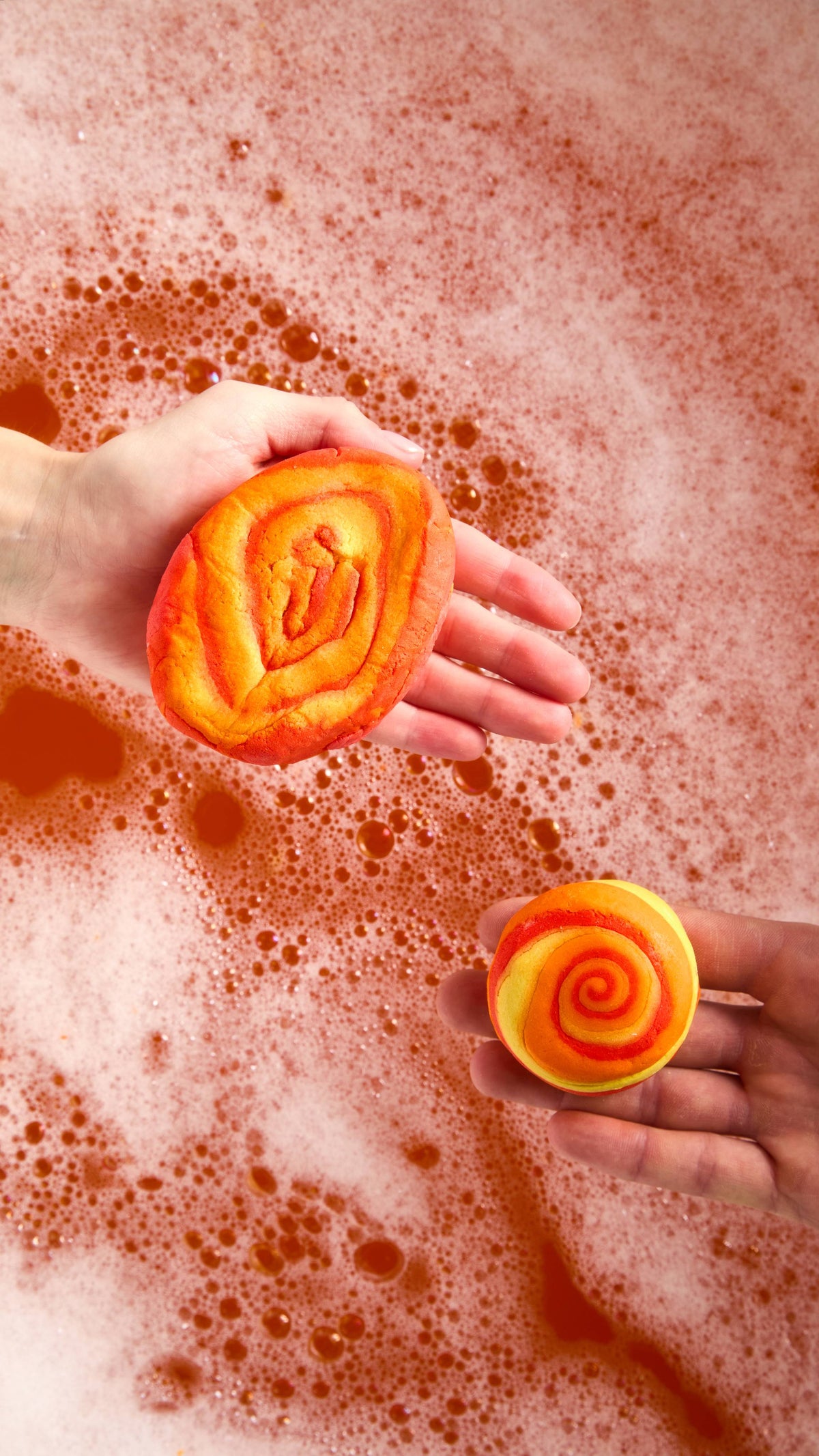 The image shows a close-up of two hands holding two different sizes of vibrant Brightside bubble bars above the firey orange, bubbly water below.