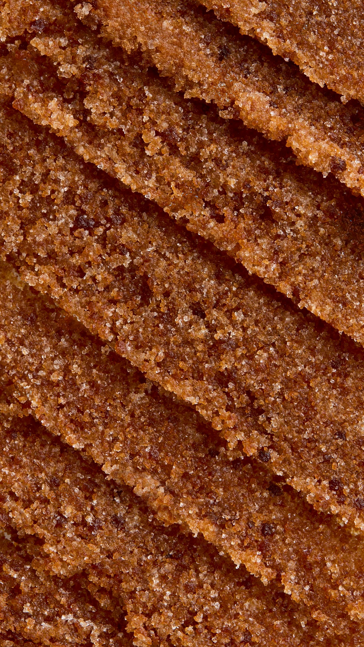 A close-up, aerial view of textured, thick caramel-coloured Buzz lip scrub. The texture is rich and gritty, arranged in several diagonal, overlapping swoops that create ridges and valleys.